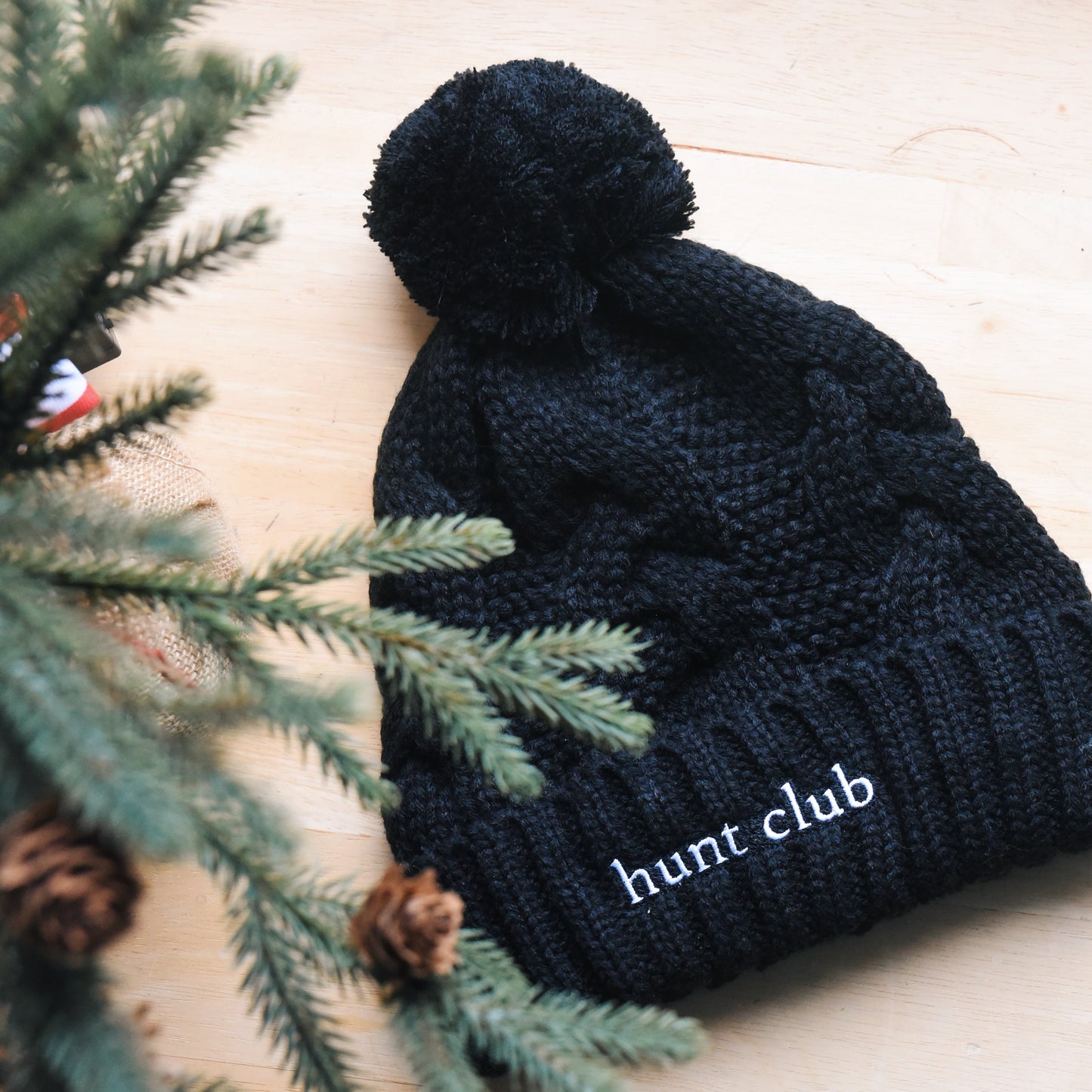 Black knit beanie with pom-pom on a wooden surface next to pine branches.